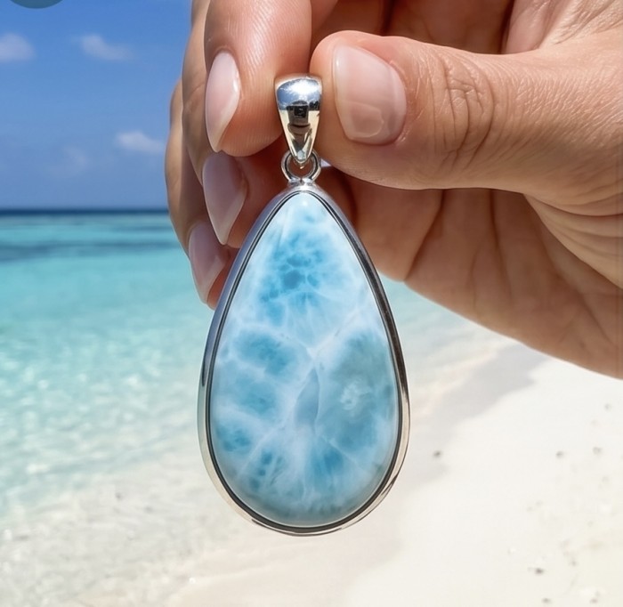 The best quality Natural Larimar from Dominican Republic .925 Sterling Silver handcrafted pendant - Close-up of a teardrop Larimar pendant held by hand on a beach background.