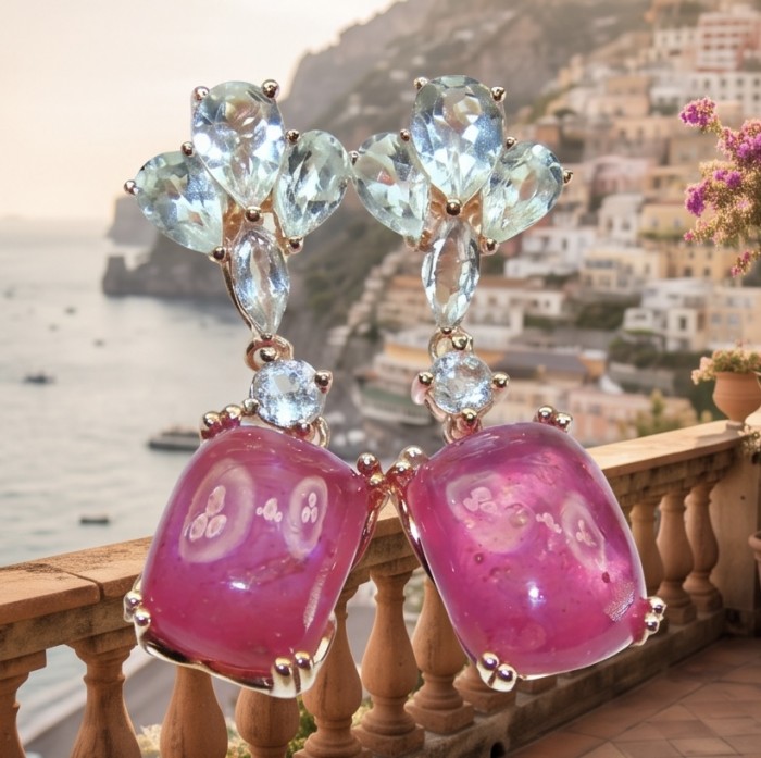 Maria Rosa - Magnificent Jewel 12.5 carat Mozambique Ruby .925 Sterling Silver dipped in 22K Rose Gold handcrafted earrings - Close-up of Maria Rosa earrings with blue and ruby gems, scenic backdrop.